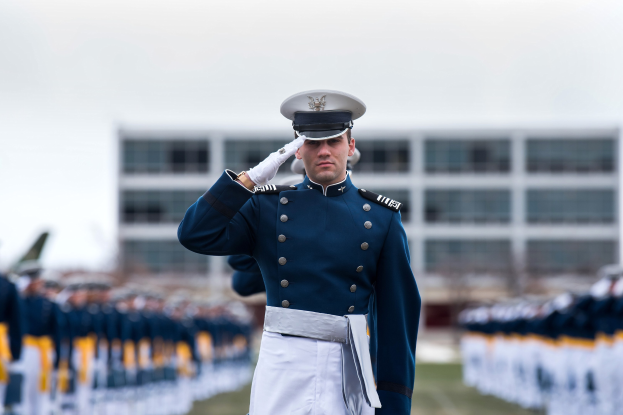 Ein Mann in einer Militäruniform salutiert auf einer Abschlussfeier und ist von einer Gruppe von Menschen umgeben, mit einem Gebäude und dem Himmel im Hintergrund.