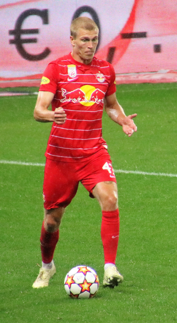 Ein Mann in roter Uniform, der auf einem Feld Fußball spielt, mit einer Tafel im Hintergrund, trägt Fußballausrüstung und -schuhe.