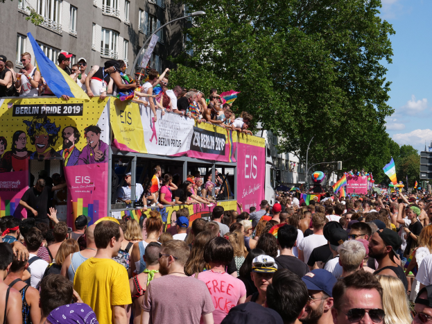 Eine gro├če Menschenmenge marschiert auf der Stra├če bei Berlin Pride 2019, viele tragen M├╝tzen und Schutzbrillen, halten Fahnen und Transparente, mit Geb├Ąuden, Laternenmasten und B├Ąumen im Hintergrund bei bew├Âlktem Himmel.
