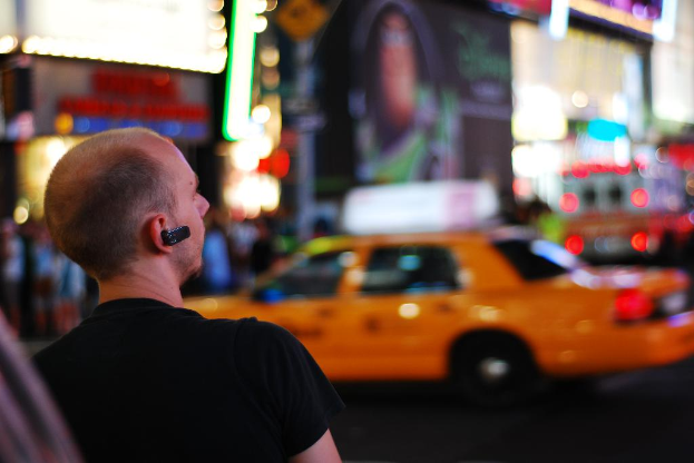 Eine Person in einem schwarzen T-Shirt und einem schwarzen Bluetooth-Gerät steht vor einem gelben Auto auf einer Straße, mit anderen unscharfen Objekten im Hintergrund.