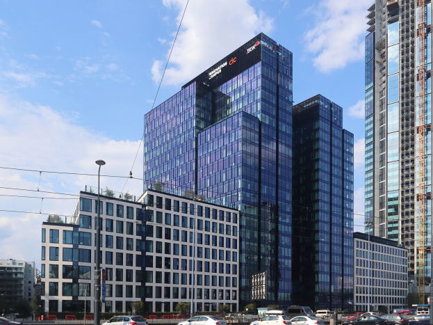 Außenansicht des Hauptsitzgebäudes der Deutschen Bank in Frankfurt, Deutschland, mit städtischer Umgebung einschließlich Straßeninfrastruktur, Fahrzeugen, Grünflächen und einem bewölkten Himmel.