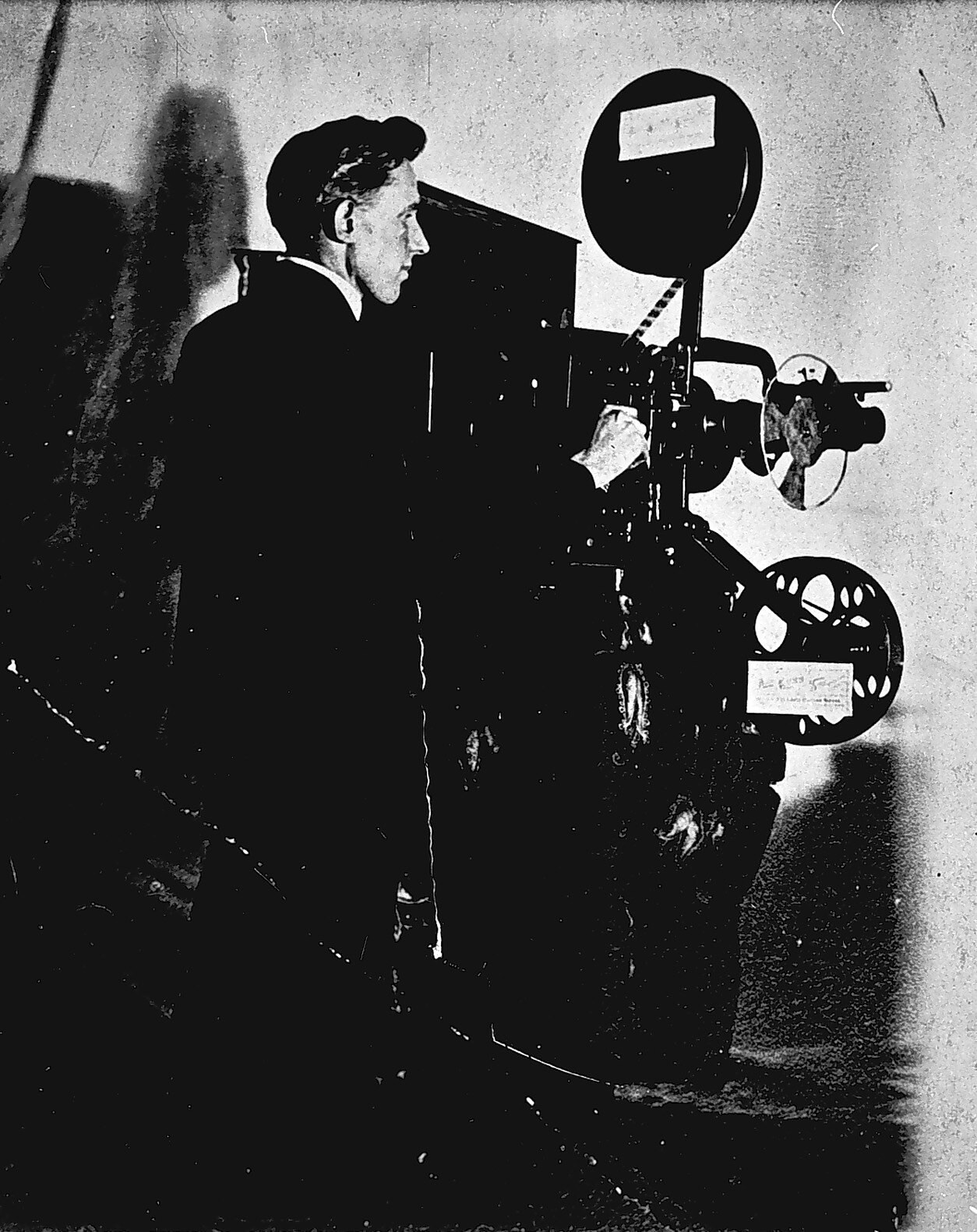 Schwarzes und weißes Foto eines Mannes in einem Anzug, der neben einem Filmprojektor steht und einen ernsten Gesichtsausdruck hat.