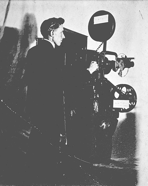 Schwarzes und weißes Foto eines Mannes in einem Anzug, der neben einem Filmprojektor steht und einen ernsten Gesichtsausdruck hat.