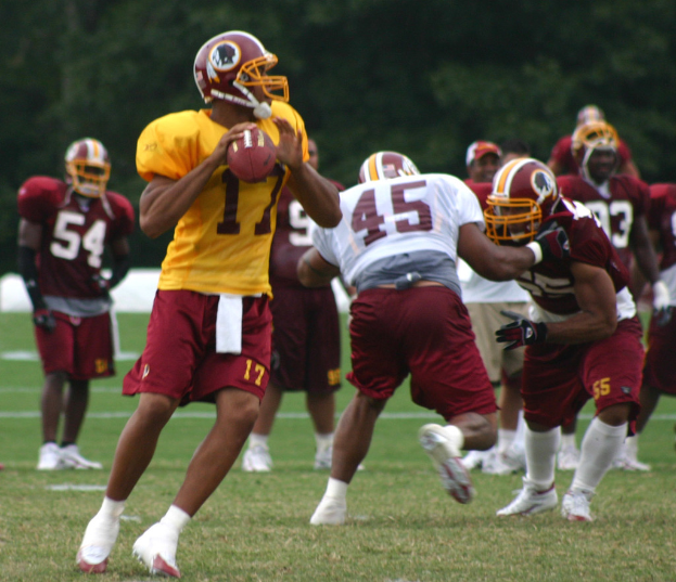 Eine Gruppe von Personen in Trikots, Helmen und weißen Schuhen spielt American Football auf einem grasbewachsenen Feld mit Bäumen im Hintergrund.