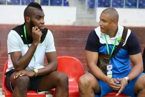 Zwei Männer sitzen auf roten Stühlen in einem Stadion, einer trägt ein weißes und schwarzes T-Shirt und blaue Shorts, der andere eine ID-Karte und scheint angeregt zu diskutieren.