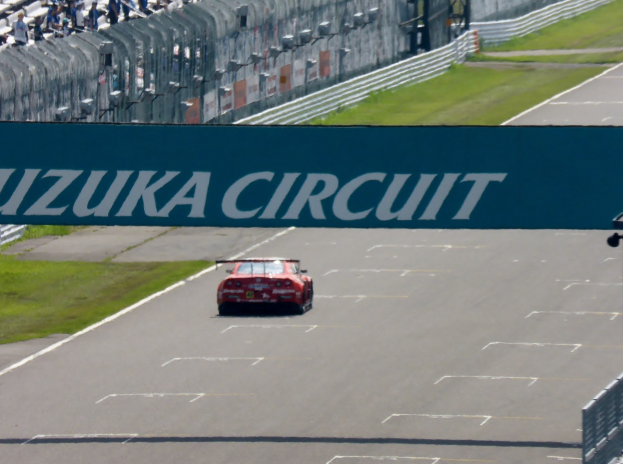 Roter Sportwagen auf einer Rennstrecke mit "Suzuka Circuit"-Schriftzug im Hintergrund, umgeben von Gras und Absperrungen, mit Zuschauern auf den Tribünen.