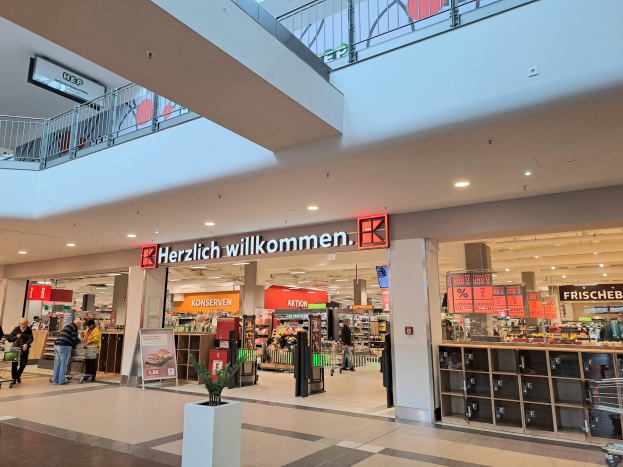 Innenansicht eines Einkaufszentrums mit Menschen, Geschäften, Infotafeln mit der Aufschrift "Herzlich willkommen", Beleuchtung, Geländern und einem Topfpalme unten.