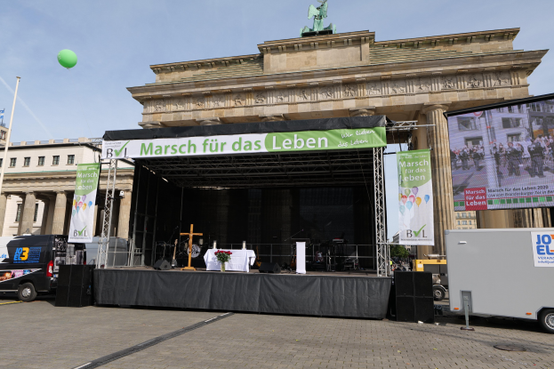 Bühne vor dem Brandenburger Tor mit einem Tisch, Bannern, Lautsprechern, Fahrzeugen, Gebäuden, einer Statue, einer Flagge und Wolken am Himmel.