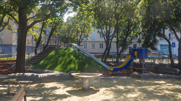 Ein Spielplatz mit einer Rutsche und einer Bank, umgeben von Bäumen, Bänken, Treppen, Geländern, Pflanzen, Gras, Steinen und Gebäuden im Hintergrund unter einem klaren blauen Himmel.
