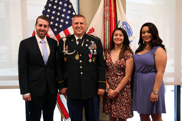 Zwei Frauen in Kleidern, eine Person in Uniform und ein Mann in Anzug stehen und lächeln drinnen, mit Jalousien und Fahnen im Hintergrund.
