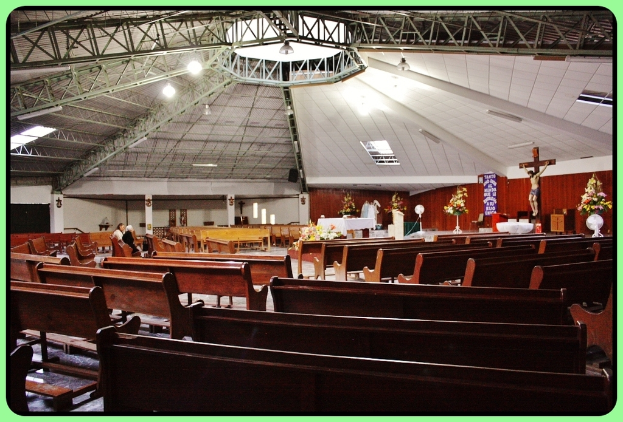 Ein Innenblick auf eine Kirche mit Holzreihenbänken, einigen Sitzenden, einer Statue und Blumensträußen im Hintergrund und einer Decke mit sichtbarer Dachkonstruktion.