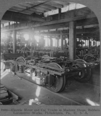 Schwarzes und weisses Foto eines Elektromotors und eines Lastwagens in einer Fabrik mit Maschinen, Saulen und einem Dach, mit Text unten.