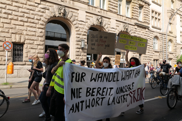 Eine Gruppe von Menschen marschiert während einer Demonstration in Berlin die Straße entlang, einige halten Schilder und Banner, andere fahren Fahrräder, im Hintergrund ein Gebäude mit Fenstern, Bögen, Säulen und Skulpturen.
