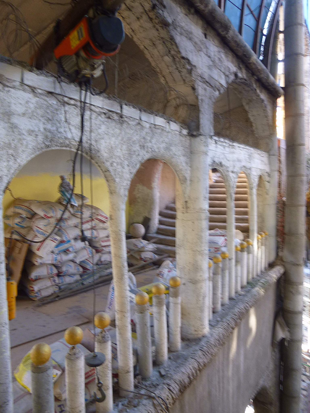 Ein altes Gebäude mit Bögen und Säulen unter Renovierung, mit verstreuten Werkzeugen, Materialien und verschiedenen Gegenständen, einschließlich Taschen, Geländern und einer Maschine mit Drähten oben.