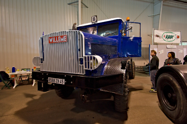 Ein Lastwagen ist in der Mitte des Bildes zu sehen, neben dem auf der rechten Seite ein paar Menschen stehen. Im Hintergrund ist eine Wand mit einem Tisch davor, auf dem ein Krug, eine Tasse und Decken stehen, umgeben von Stühlen. Auf der rechten Seite ist ein weiteres Fahrzeug mit einem sichtbaren Reifen zu sehen.