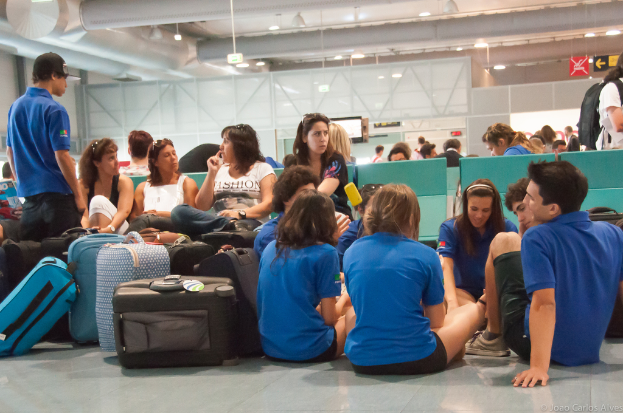 Ein belebter Flughafen-Terminal mit vielen Menschen, die auf Flüge warten, Gepäck auf der linken Seite und Glastüren mit Richtungsschildern im Hintergrund.