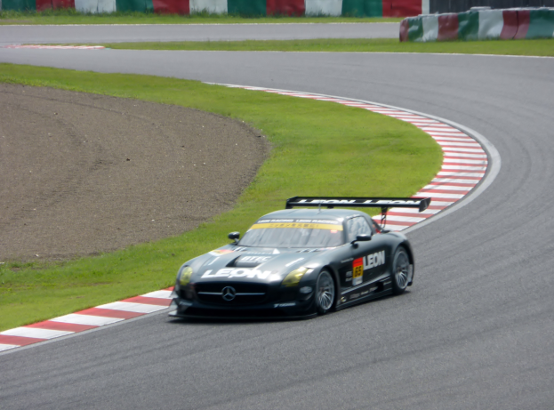 Ein Mercedes-Benz SLS AMG GT3 ist auf der Nürburgring positioniert, mit grünem Gras und einer Wand im Hintergrund und der Straße, die sich vor ihm erstreckt.