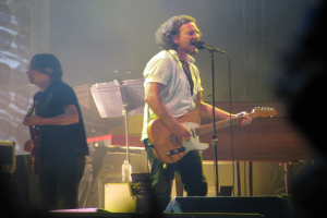 Ein Mann in einem weißen Hemd singt in ein Mikrofon und spielt Gitarre auf der Bühne, begleitet von einem anderen Mann in einem schwarzen Hemd, der ebenfalls Gitarre spielt, mit einem Piano, Kabeln, Lautsprechern und Bühnenbeleuchtung im Hintergrund.