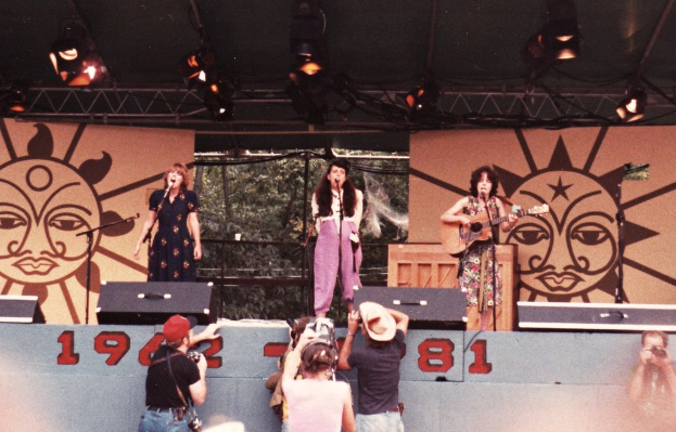 Drei Mädchen auf der Bühne, zwei singen in Mikrofone und eines spielt Gitarre, mit Zuschauern im Vordergrund, die Fotos machen, und einem braunen Bühnenrahmen mit Scheinwerfern im Hintergrund.