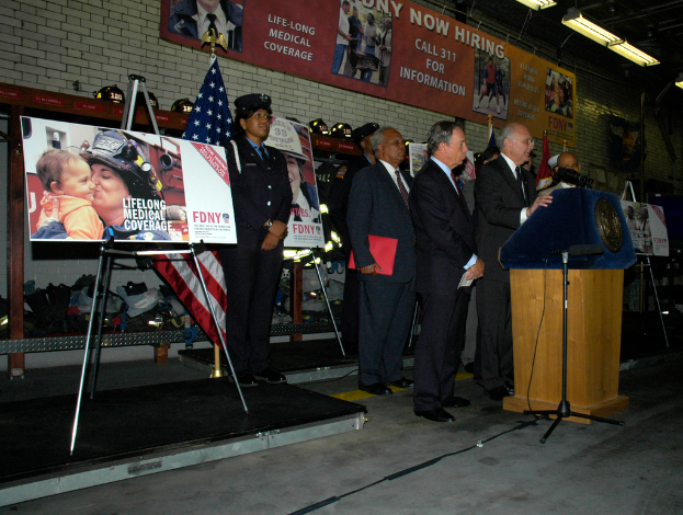 Eine Gruppe von Menschen in der Nähe eines Podiums mit Mikrofonen, Schildern auf Ständern, Flaggen und Schildern an der Wand, alles unter hellen Lichtern.