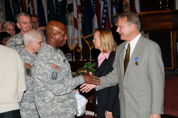 Eine Gruppe von Menschen, die stehen, mit zwei Personen im Vordergrund, die Hände schütteln, Fahnen im Hintergrund, ein Podium mit Mikrofon und eine Topfpflanze in der Nähe.