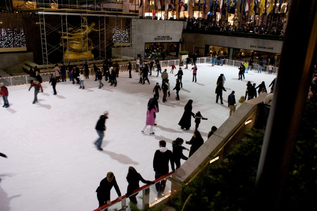 Eine Gruppe von Menschen, die auf einer zugefrorenen Fläche Schlittschuh laufen, umgeben von Gebäuden.