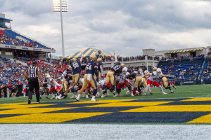 Ein Football-Spiel im Gange mit Spielern in Helmen und Sportkleidung im Vordergrund, Zuschauern, Geländern, Beschriftungen, Laternenpfählen und einem bewölkten Himmel im Hintergrund und Text am unteren Rand des Bildes.