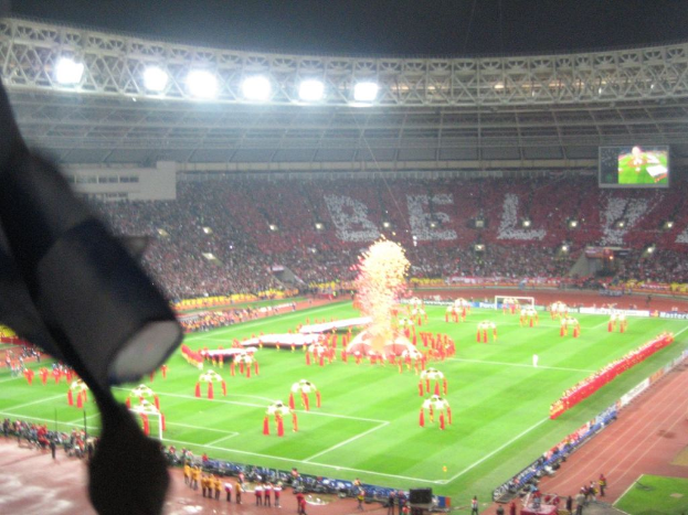 Ein Stadion mit Flutlicht, Eisengittern, Zuschauern, stehenden Individuen, Werbetafeln, Bildschirmen, Sicherheitskameras und dekorativen Fahnen.