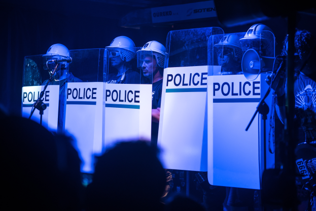 Eine Gruppe von Polizisten in Helmen steht vor einer Menge, hält Schilder mit Text, mit Mikrofonen und Ständern im Vordergrund.