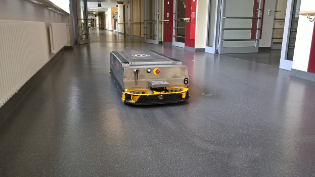 Ein Roboter zum Reinigen von Böden steht auf dem Boden eines Gebäudes, umgeben von Wänden, Türen, Lampen und anderen Gegenständen.