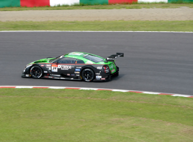 Ein roter Sportwagen mit Sponsorentext und Nummern fahrt auf einer Rennstrecke, die von Gras umgeben ist, mit einer Wand im Hintergrund w├Ąhrend der 24 Stunden von Le Mans.