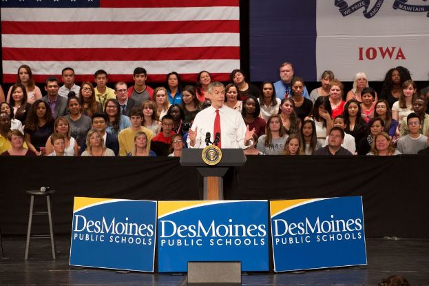 Ein Mann in einem weißen Hemd und roter Krawatte steht an einem Rednerpult mit drei Texttafeln, einem Hocker mit Mikrofon und einem sitzenden Publikum im Hintergrund, mit einer Flagge und zusätzlichem Text.