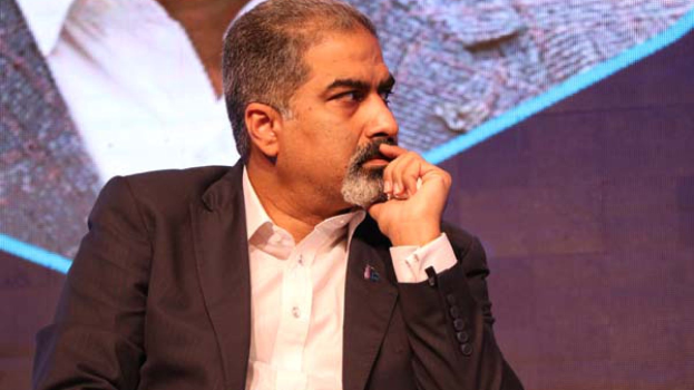 Ein Mann in Anzug sitzt auf der Bühne und denkt nach vor einem Bildschirm.