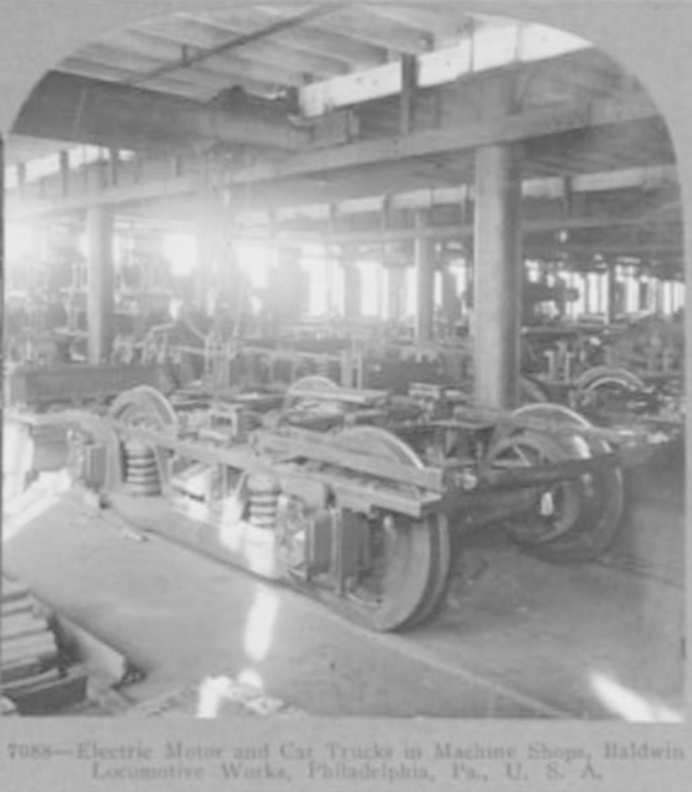Schwarzes und weißes Foto eines Elektromotors und eines Lastwagens in einer Fabrik mit Maschinen, Säulen und einem Dach, mit Text am unteren Rand.