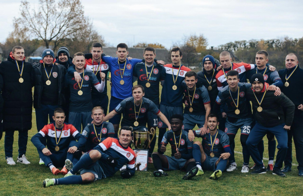 Eine Gruppe von Männern in Medaillen steht auf einem Fußballfeld und hält einen Pokal, mit Bäumen und einem klaren blauen Himmel im Hintergrund.