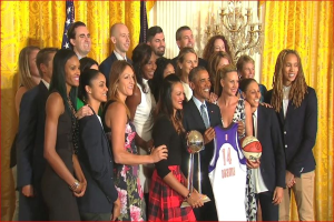 Präsident Obama und First Lady Michelle Obama posieren mit dem Frauen-Basketball-Team im Oval Office des Weißen Hauses, halten einen Basketball, Pokal und lächeln mit einer Flagge, Vorhängen und einer Kerzenleuchter im Hintergrund.