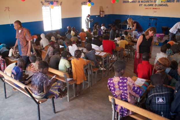 Kinder auf Bänken im Vordergrund, Erwachsene führen sie an, im Hintergrund Stühle, eine Tür, ein Fenster und ein Tisch sowie Ballons und Text oben.