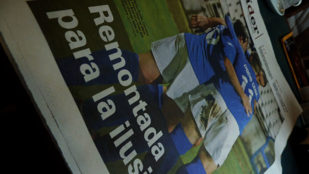 Eine Zeitung auf einem Tisch zeigt drei Männer in blauen T-Shirts, möglicherweise beim Fußballspielen, mit Text unten im Bild.