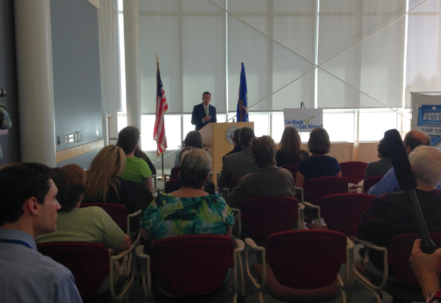 Ein Mann steht an einem Podium und spricht zu einem Publikum, das auf Stühlen sitzt, mit zwei Fahnen daneben und einer Wand, Säulen und Fenstern im Hintergrund.