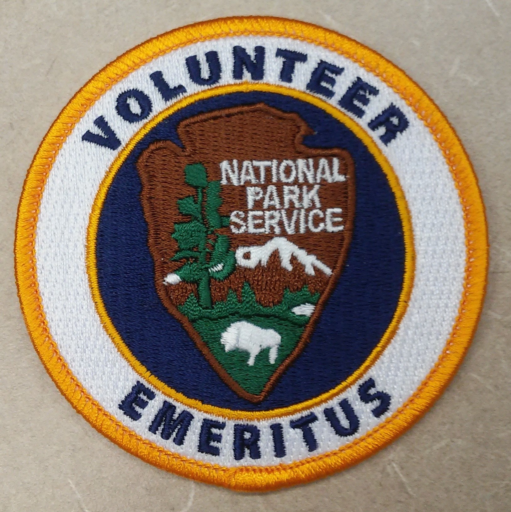Runder Flicken mit blauem Rand und weißem Hintergrund, der schwarzen Text "Volunteer National Park Service Emeritus" in der Mitte eines gelben Kreises zeigt.