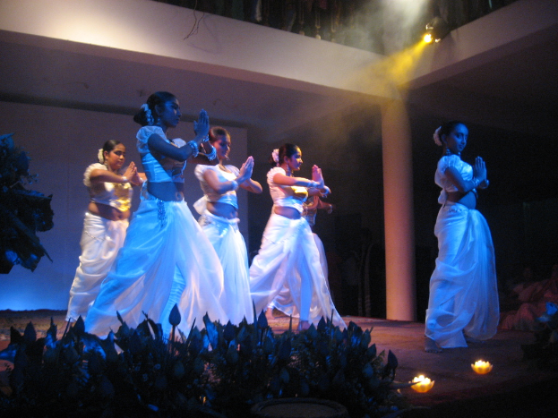 Eine Gruppe von Menschen tanzt auf einer Bühne mit Pflanzen davor und einer Zuschauermenge.