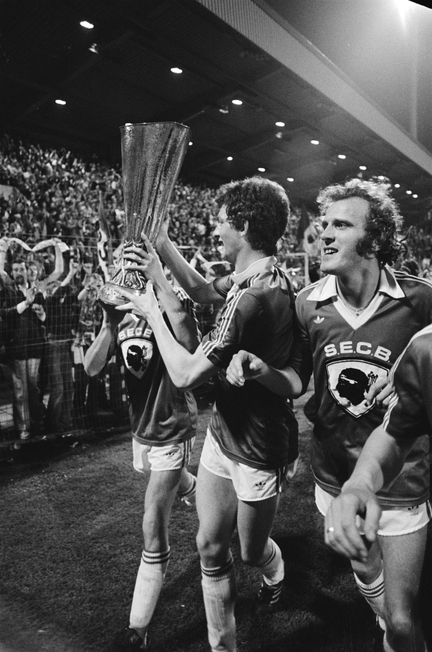 Schwarzes und weißes Bild von Fußballspielern, die einen Pokal auf einem Feld halten und feiern, mit Zuschauern hinter einem Zaun unter Stadionbeleuchtung.