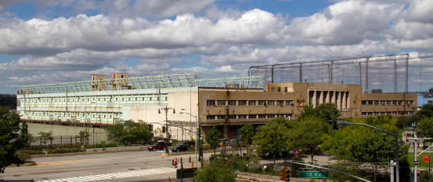 Eine Straße mit Fahrzeugen, Bäumen und Straßenlaternen neben einer Wasserfläche, mit einem großen Gebäude im Hintergrund.