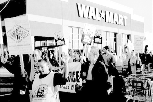 Eine Gruppe von Menschen protestiert vor einem Einkaufszentrum und hält Plakate in den Händen, dargestellt in Schwarz-Weiß.