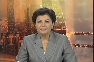 Eine Frau in einem Business-Outfit sitzt vor einem Fernseher, mit ein paar Gebäuden im Hintergrund, die scheinbar eine Nachrichtensprecherin zeigt, wie der Text am unteren Bildschirmrand anzeigt.