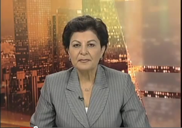 Eine Frau in einem Business-Outfit sitzt vor einem Fernseher, mit ein paar Gebäuden im Hintergrund, die scheinbar eine Nachrichtensprecherin zeigt, wie der Text am unteren Bildschirmrand anzeigt.