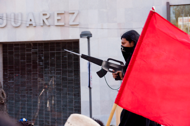 Ein Mann in einem schwarzen Kleid hält eine Waffe und steht neben einer roten Fahne, mit einer Straßenlaterne in der Nähe einer Wand und einem Gebäude im Hintergrund, und lesbarer Text in der oberen linken Ecke auf der Wand.