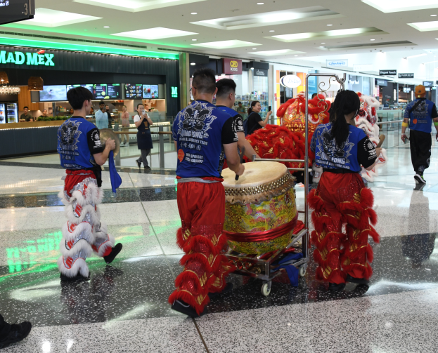 Eine Gruppe von Menschen in roter und blauer Kleidung durchquert einen Flughafen, einige tragen Musikinstrumente und Gepäckwagen, mit Geschäften, Namensschildern und Deckenleuchten im Hintergrund, was auf eine chinesische Neujahrsfeier in Singapur hindeutet.