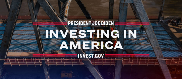 Präsident Joe Biden in der Nähe einer Brücke mit dem Text "Investing in America" und verstreuten Gegenständen im Vordergrund.