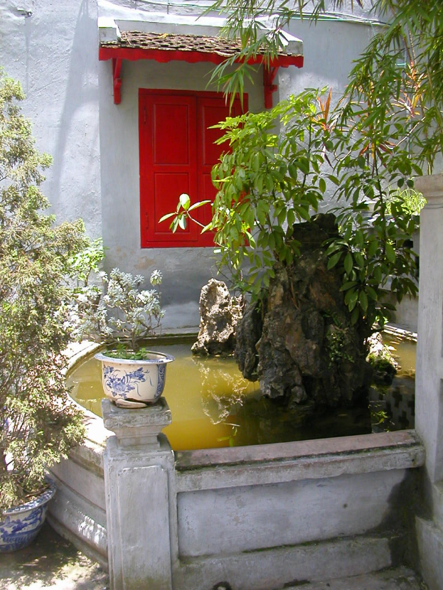 Kleiner Hof mit einer zentralen roten Tür, umgeben von Bäumen, Pflanzen, Felsen, einer Wand und einem Topf mit einer Pflanze.